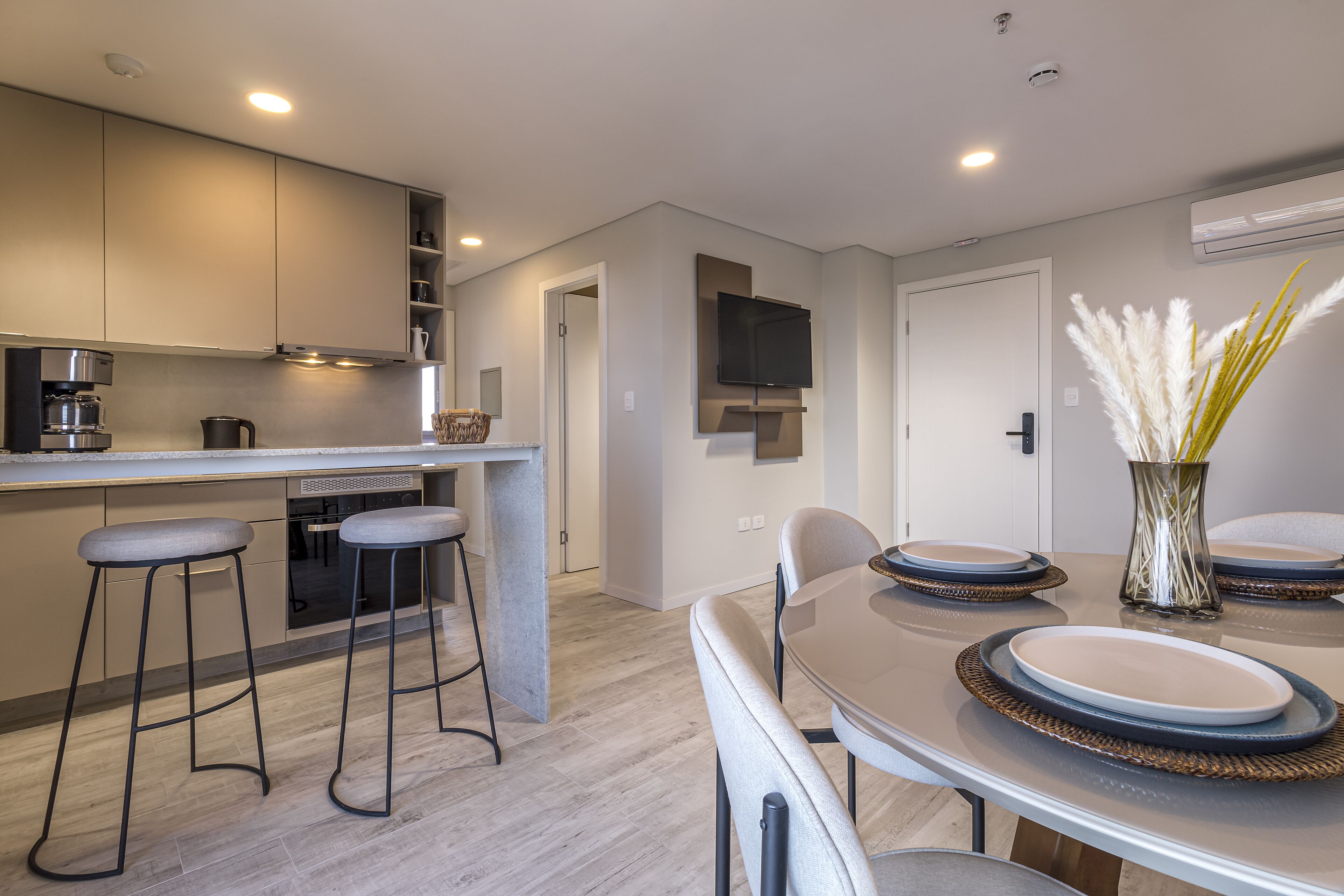 A modern kitchen with a dining area, featuring a built-in oven, a coffee machine, and a flat-screen TV.