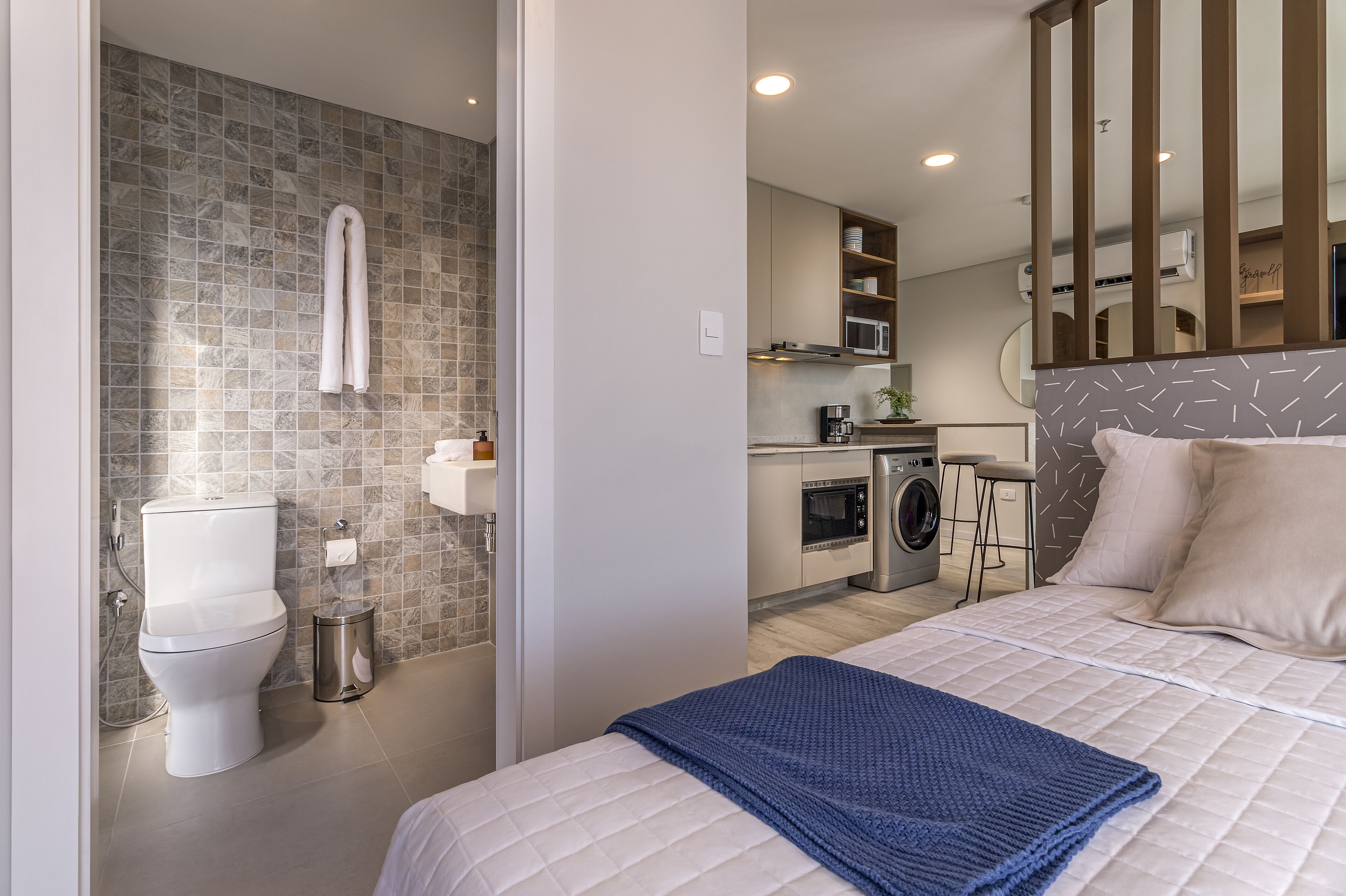 Une chambre d’hôtel moderne avec salle de bain, kitchenette et un lit recouvert d’une couverture bleue.