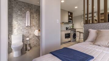 Une chambre d’hôtel moderne avec salle de bain, kitchenette et un lit recouvert d’une couverture bleue.