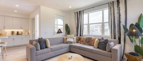 A modern living room with a grey sectional sofa, a round coffee table, and large windows with patterned curtains.