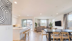 A modern kitchen with a central island, a dining area with a large table and chairs, a living room with a sofa and TV, and a view of a residential area.