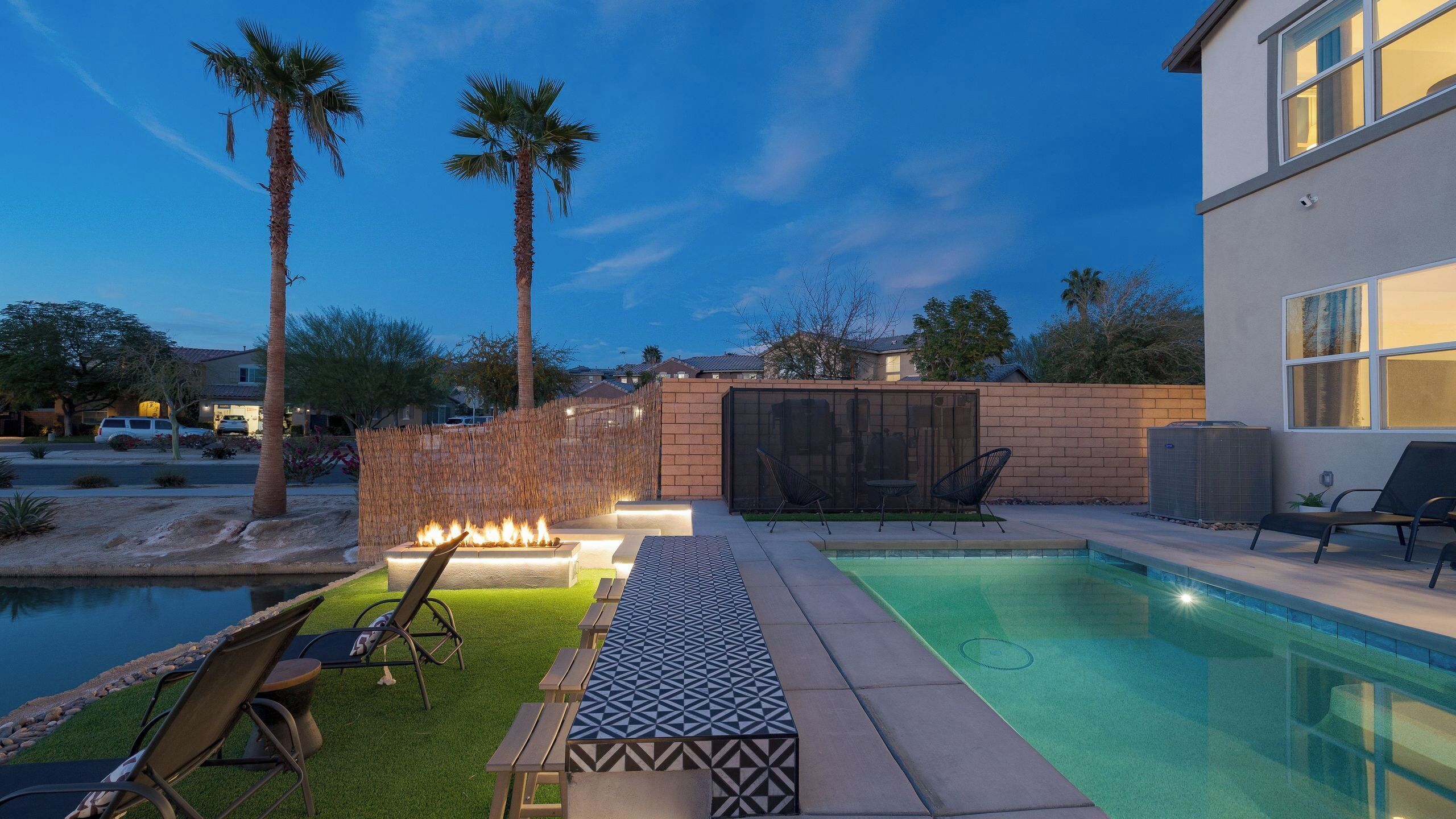 A backyard with a swimming pool, lounge chairs, a fire pit, and a modern house.