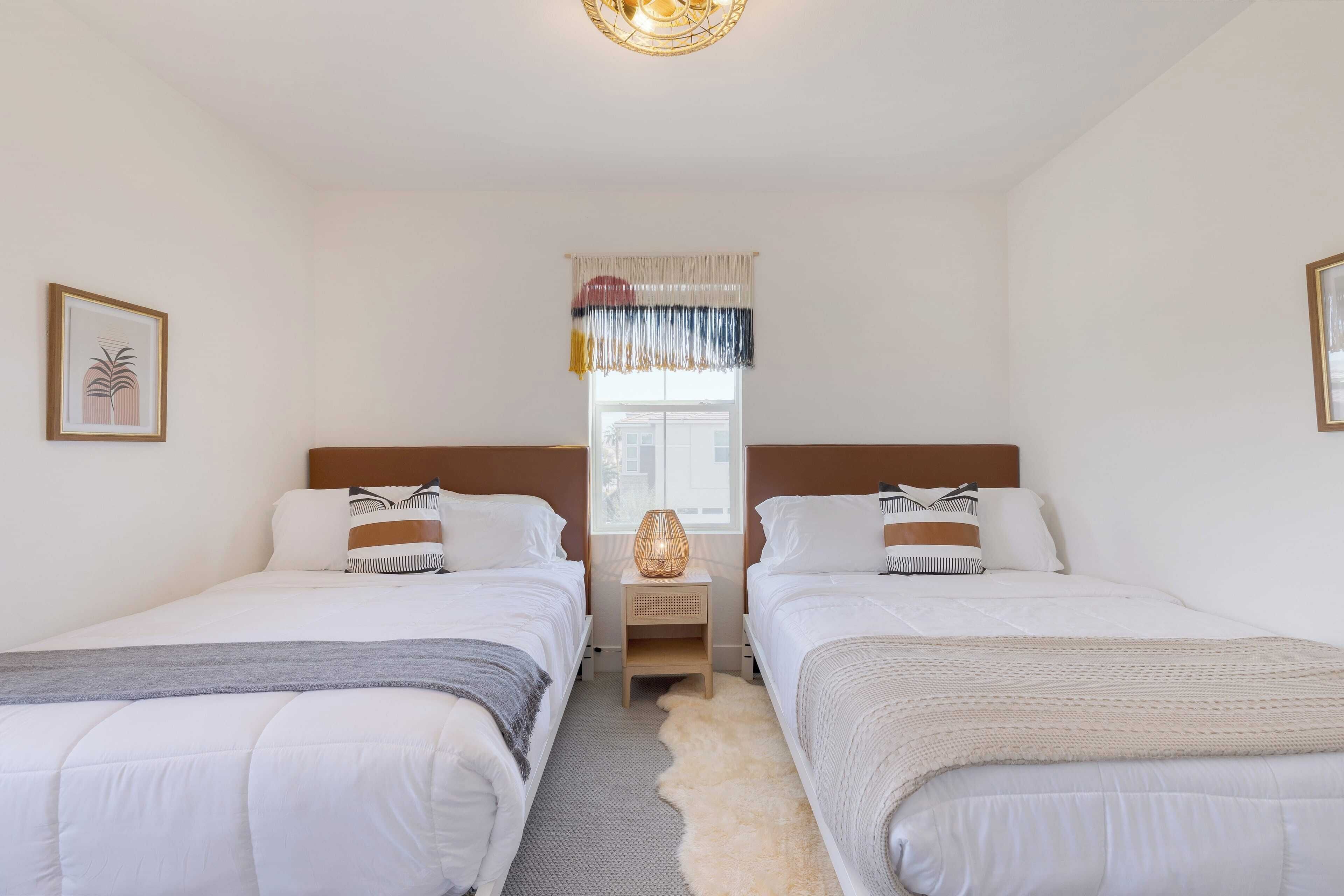 A bedroom with two beds, a window with a curtain, a nightstand with a lamp, and a framed picture on the wall.