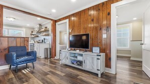 A modern living room with a TV, wooden panel walls, a blue sofa, and a kitchen in the background. - Charming farmhouse in tranquil Candler, pet friendly w/fully fenced yard (Candler)