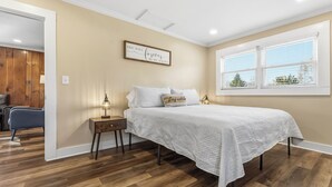 A bedroom with a bed, bedside tables, a nightstand, a window with blinds, and a door leading to another room. - Charming farmhouse in tranquil Candler, pet friendly w/fully fenced yard (Candler)