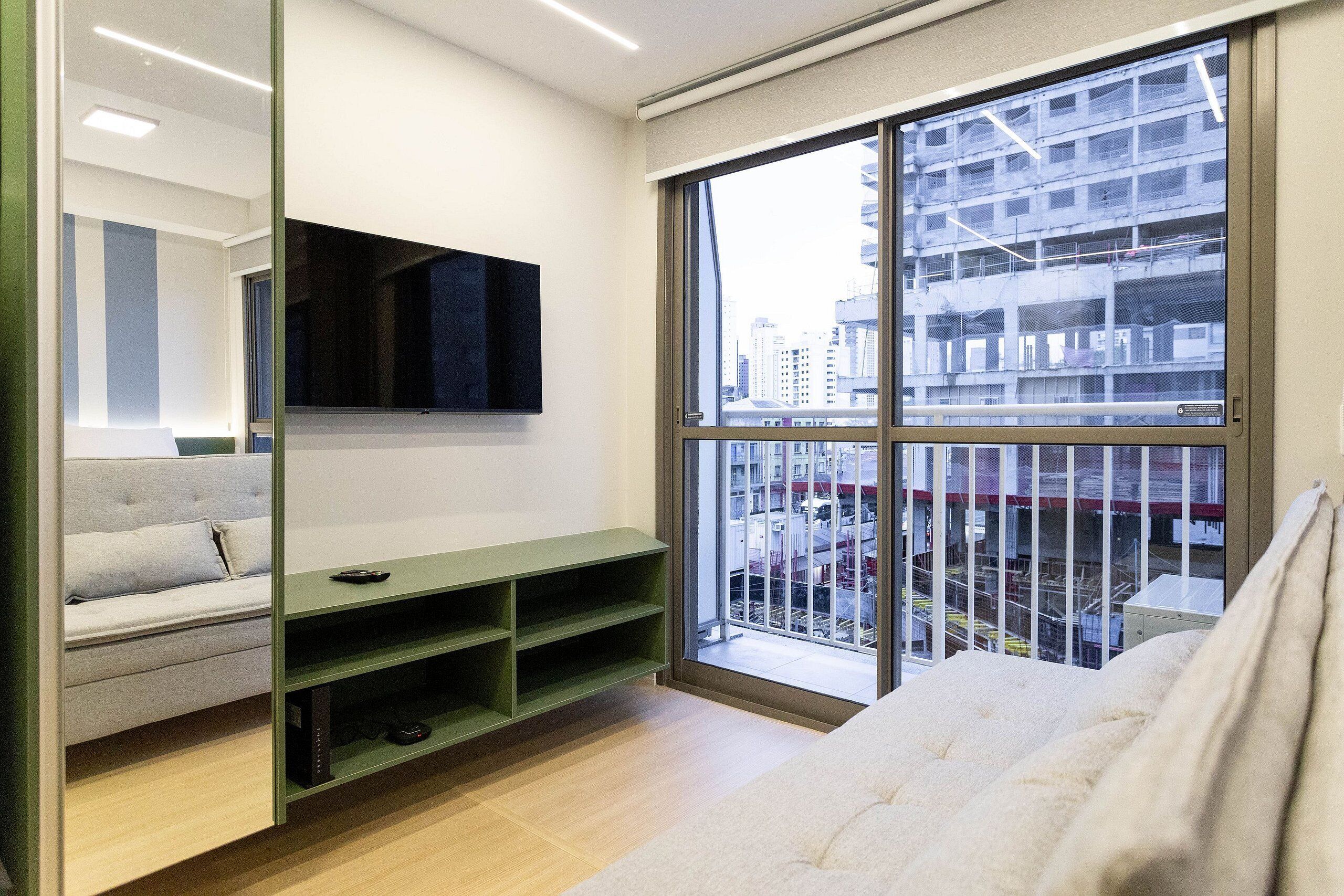 A modern hotel room with a large window, a flat-screen TV, a green console table, a sofa, and a white bed.