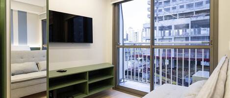 A modern hotel room with a large window, a flat-screen TV, a green console table, a sofa, and a white bed.