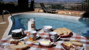 Ontbijt bij het zwembad met een geruit tafelkleed, een koffiepot, twee mokken, een mand met brood en schotels met kaas en vlees.