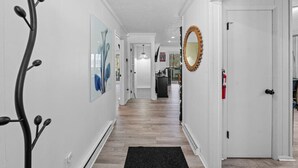 A hallway with white walls, a black rug, and a wooden floor. There are framed pictures on the walls and a mirror on the right.