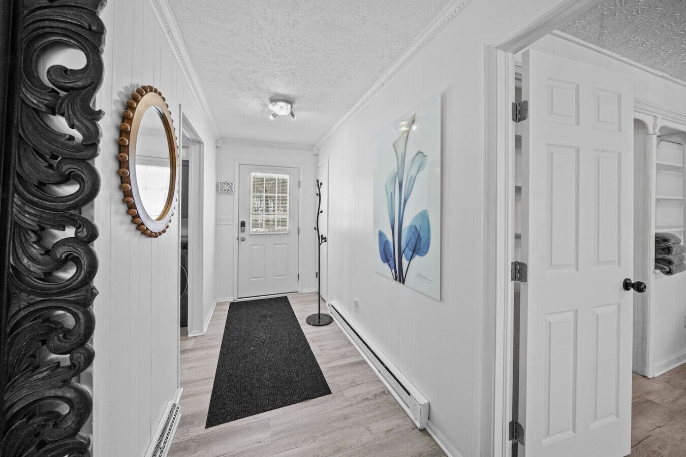 A hallway with white walls, a black floor mat, a door with a white frame, and a wall-mounted mirror.
