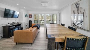 A spacious living room with a long wooden dining table, a leather sofa, and a flat-screen TV mounted on the wall.