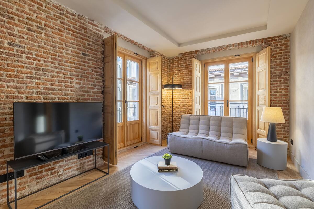 A living room with a brick wall, a flat-screen TV, a grey sofa, a round coffee table, and a lamp.