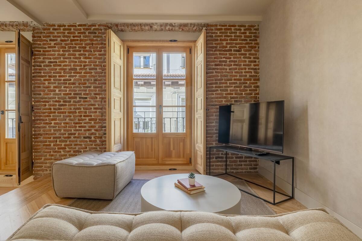 A living room with a brick wall, a wooden door, a flat-screen TV on a stand, a round coffee table with books and a plant, and a beige sofa.