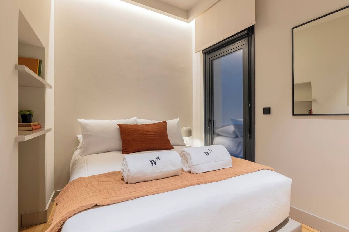 A neatly made bed with white linens, a brown throw blanket, and two towels with a logo. There is a shelf with books and a plant, and a large mirror on the wall.
