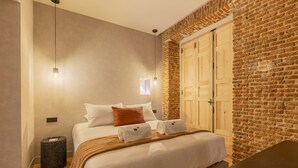 A hotel room with a brick wall, a bed with white bedding, a small trash bin, and a wooden desk. - Rio by Wynwood House (Madrid)