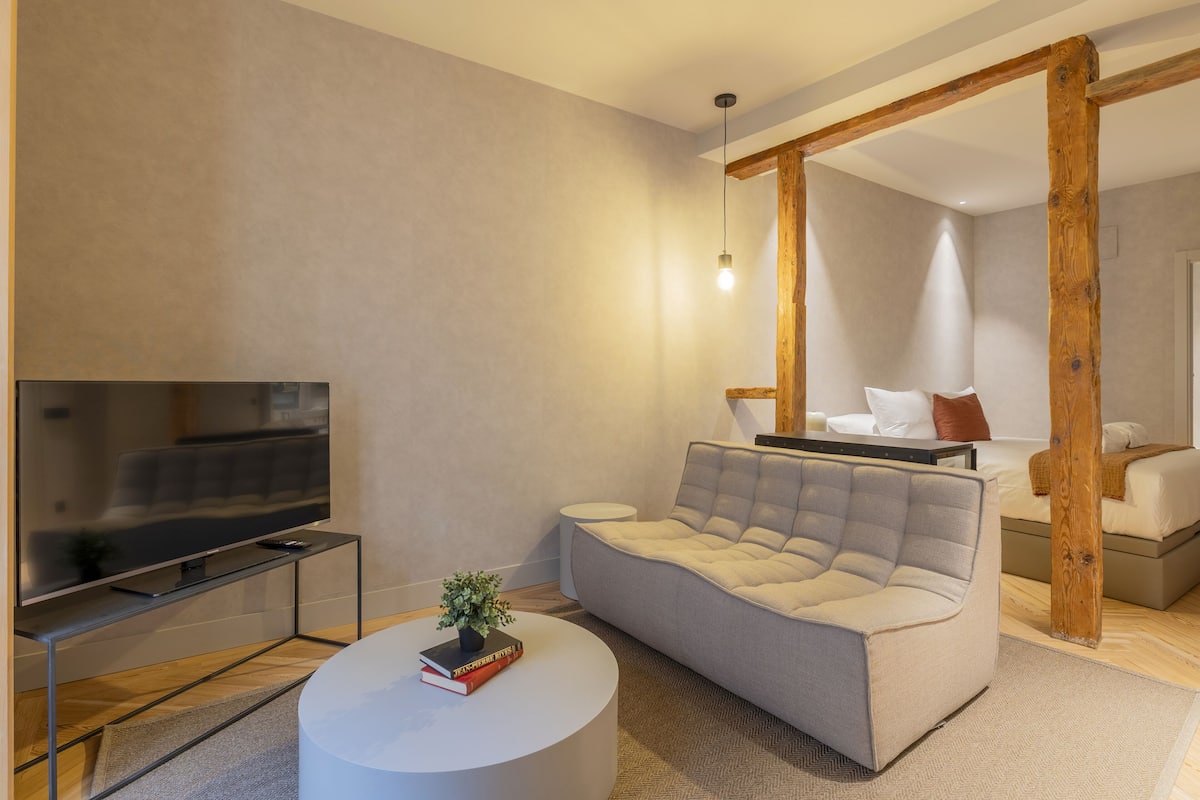 A modern living room with a flat-screen TV, a round coffee table with books, a grey sofa, and a wooden beam structure.