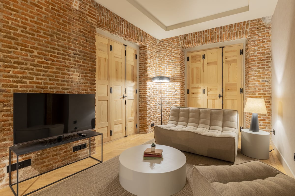 A modern living room with a brick wall, a flat-screen TV, a beige sofa, a round coffee table, and a floor lamp.