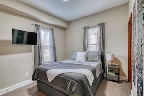 A bedroom with a bed, a TV mounted on the wall, a nightstand with a lamp, and a window with curtains.