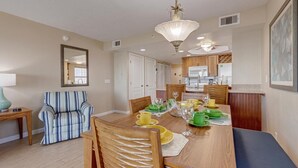 A dining area with a wooden table set for four, surrounded by chairs. There is a striped armchair and a small table with a lamp. The room has a ceiling fan and a chandelier. - Cocoa Beach 2 BR 2 Bath Condo Ocean View & Balcony (Cocoa Beach)
