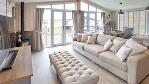 Een ruime woonkamer met een beige bank, een bijpassend poefje en een houten salontafel. Grote glazen deuren leiden naar een buitenruimte.