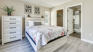 Une chambre à coucher comprenant un lit, une commode, un ventilateur de plafond et une porte donnant sur une salle de bain.