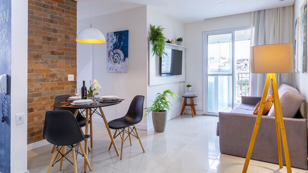 Un salon moderne comprenant un canapé, une table à manger avec des chaises, un téléviseur fixé au mur et une grande fenêtre donnant sur la ville.
