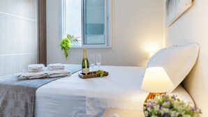 Une chambre d’hôtel avec un lit, une table de chevet avec une lampe, une fenêtre et un plateau avec une bouteille et des verres.