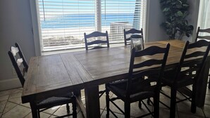 A dining area with a wooden table and chairs, large windows with blinds, and a view of the ocean.