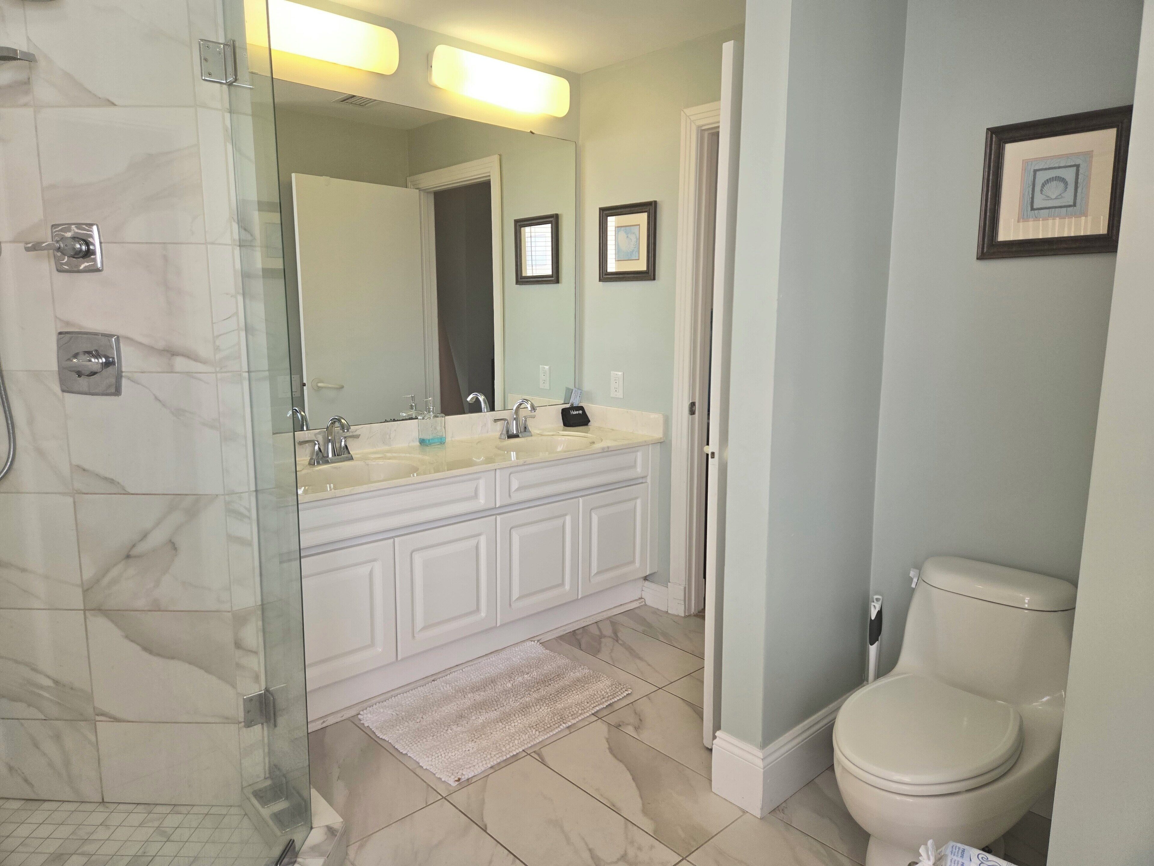 A bathroom with a glass shower enclosure, a double sink vanity, a toilet, and a framed picture on the wall.