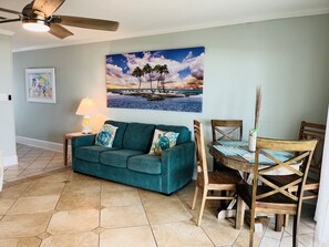 A living room with a teal sofa, a dining table with chairs, a ceiling fan, and a beach-themed painting on the wall.