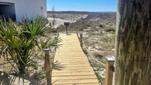 A wooden pathway leading to a modern building with a glass balcony, surrounded by dry vegetation and a clear sky. - Boardwalk #3: Beachfront + Free Daily Admissions | Pet Friendly (Pensacola)