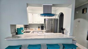 A modern kitchen with white cabinets, a large island with bar stools, stainless steel appliances, and a blue ceiling light. - Boardwalk #3: Beachfront + Free Daily Admissions | Pet Friendly (Pensacola)