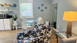 A living room with a round glass table, white wicker chairs, a blue armchair with star patterns, a yellow lamp, and a large mirror. - 3BR Coastal Cottage | Deck + Grill (Pensacola)
