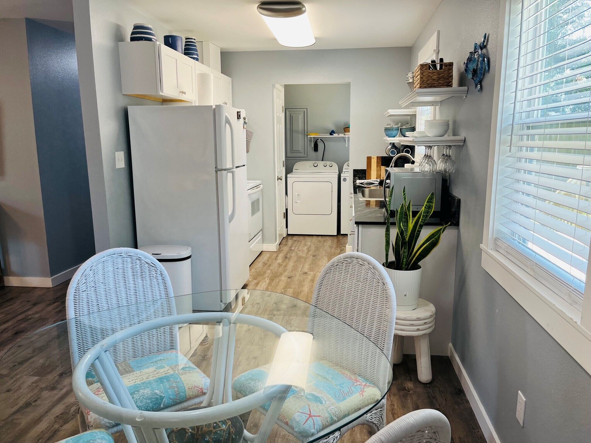 A compact kitchen with white appliances, a dining area with wicker chairs, and a potted plant.