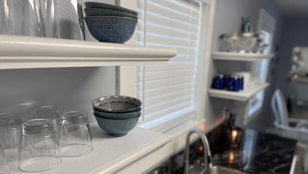 A kitchen with white shelves holding glassware and bowls, a stainless steel sink, and a window with blinds. - 3BR Coastal Cottage | Deck + Grill (Pensacola)