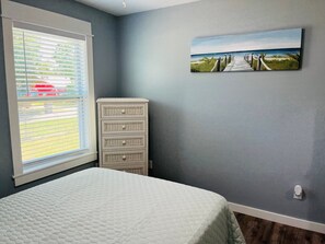A bedroom with a bed, a window with blinds, a wall-mounted painting, and a cabinet.