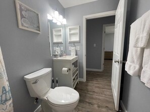 A bathroom with a toilet, sink, and mirrors. There is a towel rack and a door leading to another room. - 3BR Coastal Cottage | Deck + Grill (Pensacola)
