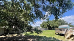 A backyard with a wooden deck, a small shed, and a grassy area with trees. - 3BR Coastal Cottage | Deck + Grill (Pensacola)
