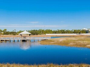 A tranquil waterfront area with a dock, a covered pavilion, and a fenced area with buildings in the background. - Grand Caribbean 214W: Perdido Key 1BR: Ocean View, Pool, Hot Tub (Pensacola)