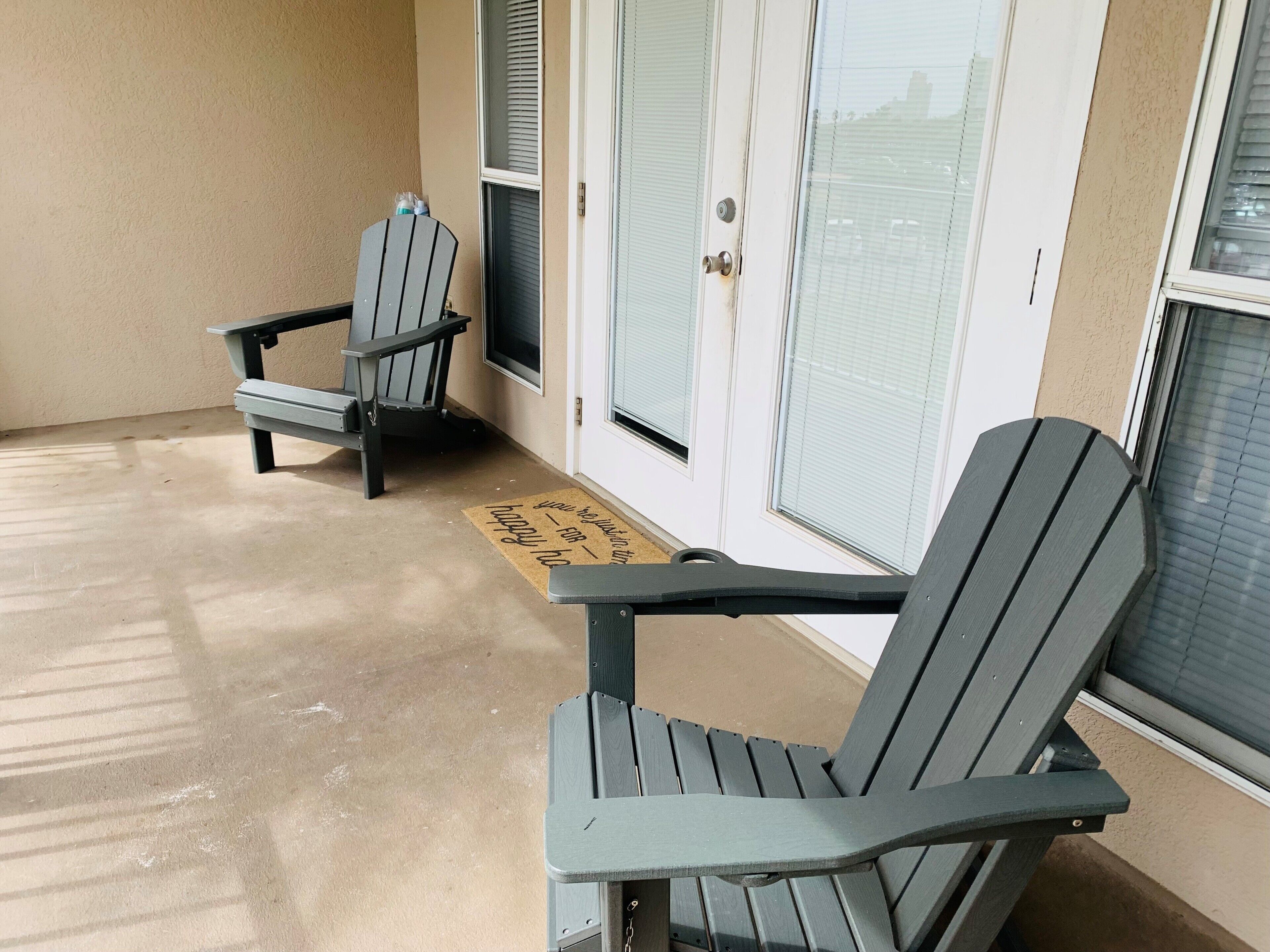 Ein Balkon mit zwei Adirondack-Stühlen, einer Fußmatte und einer weißen Tür mit Jalousien.