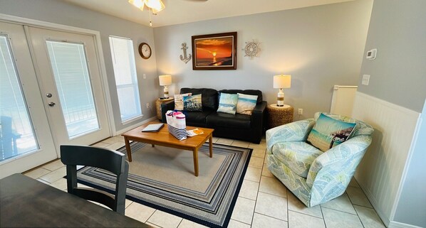A living room with a sofa, a coffee table, a chair, and a patterned armchair. There is a painting on the wall and a clock. - Grand Caribbean 214W: Perdido Key 1BR: Ocean View, Pool, Hot Tub (Pensacola)