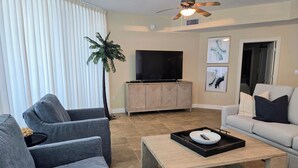 Un salon avec un canapé gris, un canapé blanc, une table basse, un téléviseur posé sur un meuble et un ventilateur de plafond.