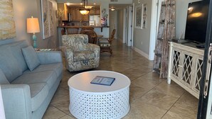 A living room with a sofa, armchair, and round coffee table. There is a television on a cabinet, and a kitchen area is visible in the background. - Ocean Breeze 602W: Gulf-Front Perdido Key 2BR/2BA + Pool (Pensacola)