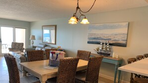 A dining area with a wooden table and woven chairs, a painting on the wall, and a ship model on a side table. - SK511: Gulf-Front 3BR Corner Condo w/ Balcony (Pensacola)