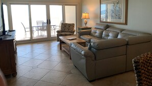 A living room with a leather sofa, a wooden coffee table, a patterned armchair, and a lamp on a side table. There is a sliding glass door leading to an outdoor area with a table and chairs. - SK511: Gulf-Front 3BR Corner Condo w/ Balcony (Pensacola)