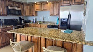 A kitchen with granite countertops, wooden cabinets, stainless steel appliances, and two bar stools. - SeaSpray 1006 Gulf Front Penthouse,Pool,Tennis (Pensacola)