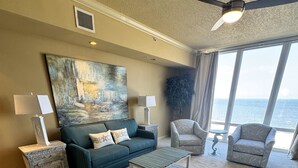 A spacious living room with a large window offering a view of the sea, a ceiling fan, and a modern sofa with matching side tables.