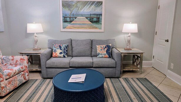 A living room with a grey sofa, two matching side tables, a round blue coffee table, a striped rug, and a framed seascape painting.