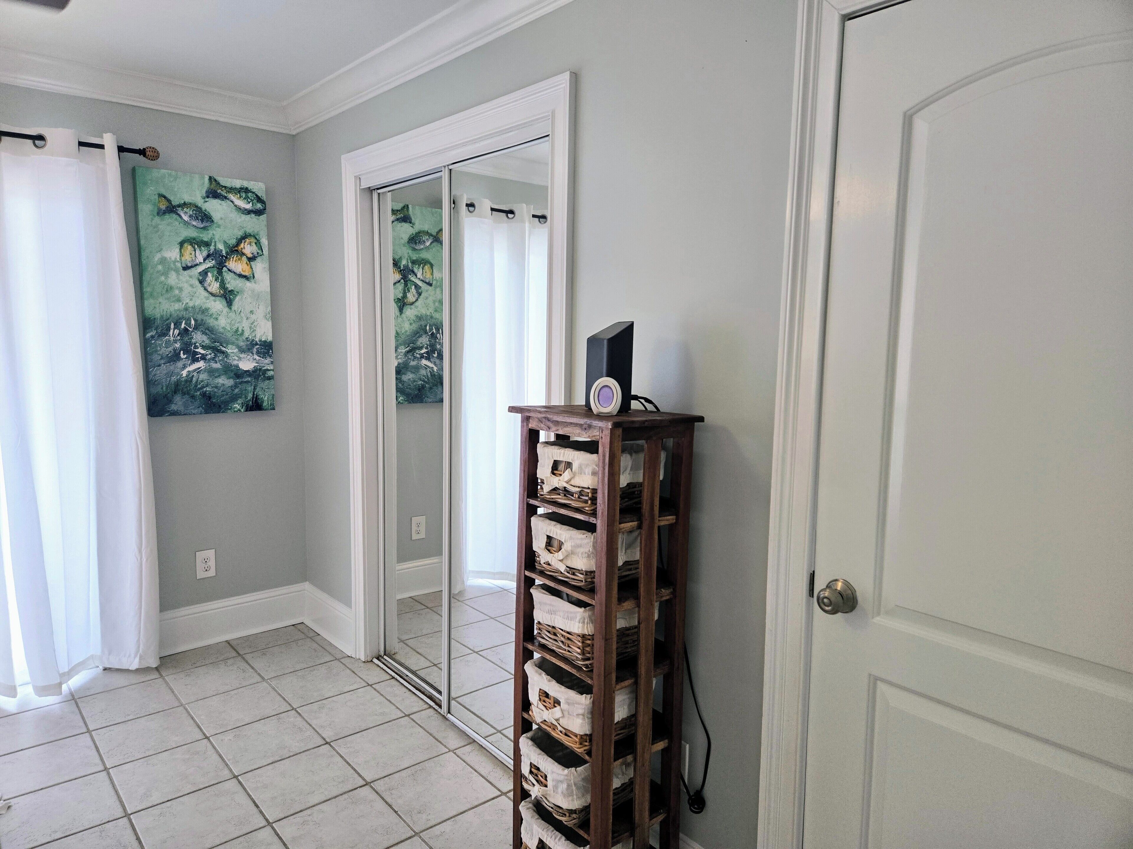 A hallway with a tiled floor, a white door, a mirror, a wooden shelf with baskets, and a painting on the wall.