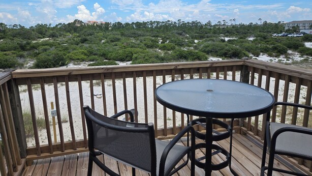A wooden deck with a round table and two chairs, overlooking a beach with sand and sparse vegetation.